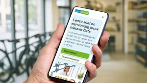 Close-up van een hand met een telefoon waarop het ontwerp van de homepagina van LeaseGemak te zien is. Op de achtergrond is het interieur van een fietsenwinkel te zien.